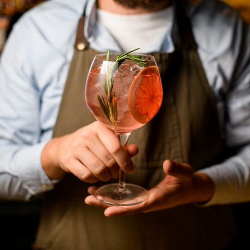 Hombre con delantal sosteniendo un servicio de ginebra con guarnición de naranja en un vaso grande con hielo y romero. 