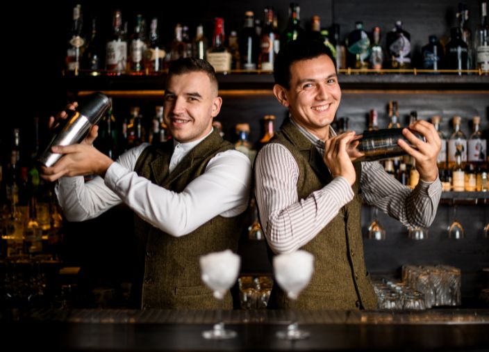 Dos bartenders hombro con hombro, sonriendo y agitando cócteles.