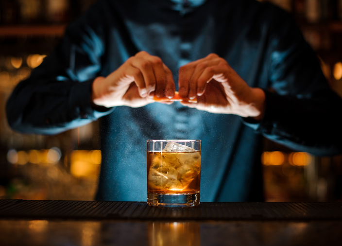 Manos exprimiendo una naranja en un cóctel de whisky.