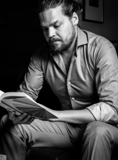 Black and white image of Zdenek Katanek reading a book sitting on a chair.