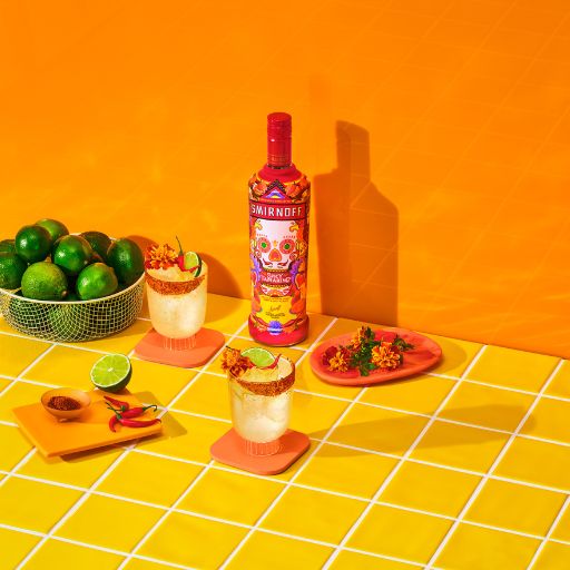 A bottle of Smirnoff Spicy Tamarind beside a cocktail, plate of spicy food and bowl of limes against an orange background. 