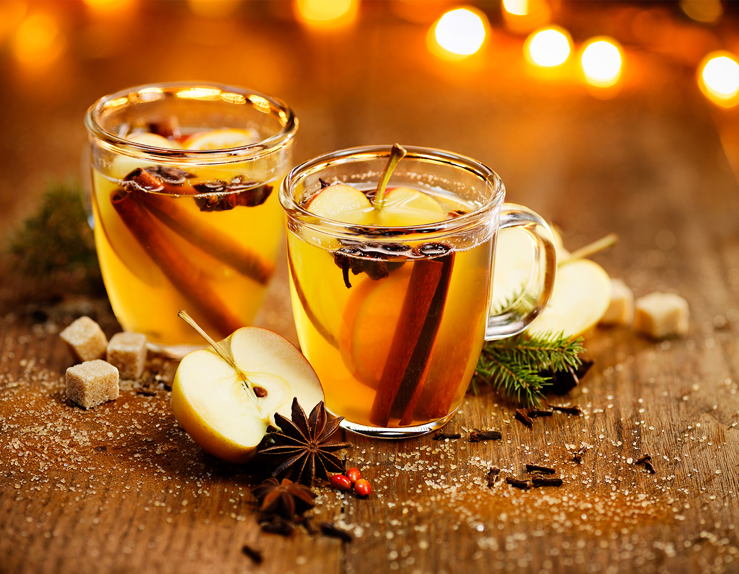 Two Toffee Apple Toddy cocktails in glass cups garnished with apple slices, a cinnamon stick and whole star anise. All surrounded by more garnish and a sprig of holly, sugar cubes and fairy lights.
