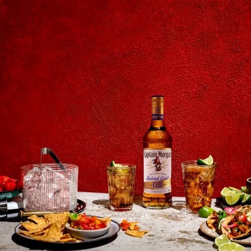 Bottle of Captain Morgan Spiced Gold 0.0% Alcohol Free Spirit on table surrounded by assorted foods and drinks against a red background