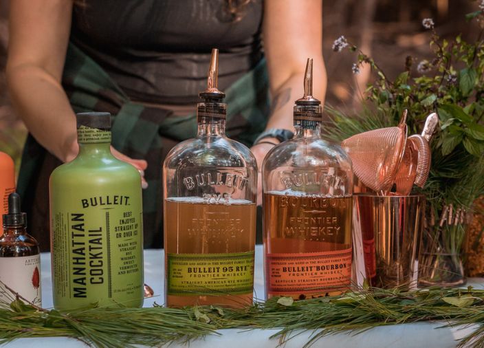 Bulleit_Bourbon_bottles_and_cocktail_with_cocktail_shaker_strainer_and_plants_on_table
