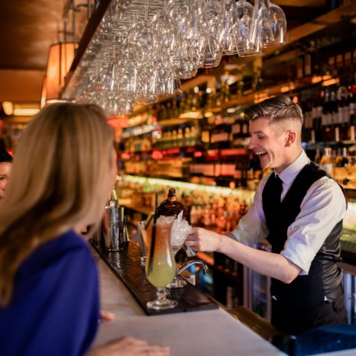  Bartender standing behind bar making cocktail whilst talking to smiling guests 