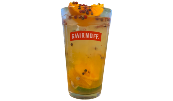 Copo alto com logo vermelho da Smirnoff contendo um coquetel refrescante amarelo-alaranjado com gelo. A bebida traz pedaços de laranja e limão mergulhados, salpicada com grãos de pimenta rosa espalhados até o topo. Fundo branco isolado.