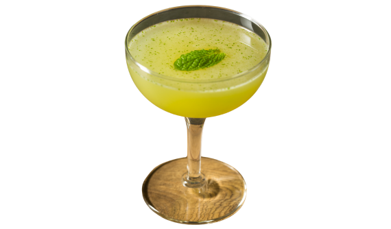 Taça coupe elegante contendo um coquetel opaco em tom verde-claro. A bebida é delicadamente decorada com uma única folha de hortelã fresca flutuando bem ao centro, cercada por minúsculos fragmentos verdes. Fundo branco isolado.