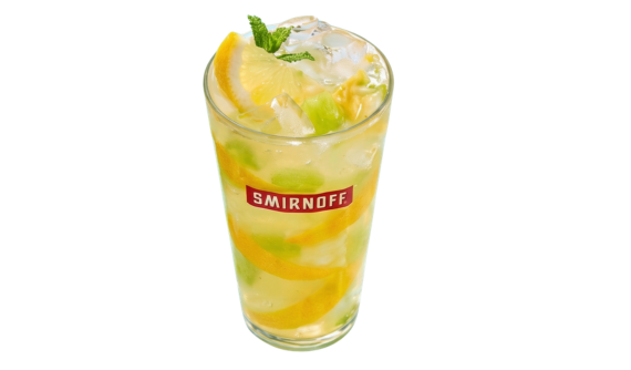 Copo alto com logo vermelho da Smirnoff contendo um coquetel amarelo-claro com muito gelo, fatias de limão siciliano e pedaços de frutas verdes. A bebida é decorada no topo com uma fatia de limão e hortelã fresca. Fundo branco.