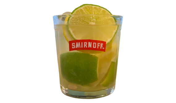 Copo baixo com logo vermelho da Smirnoff contendo um coquetel refrescante com gelo e pedaços de limão verde e siciliano. Fatias de ambos os limões decoram a borda superior. Fundo branco.