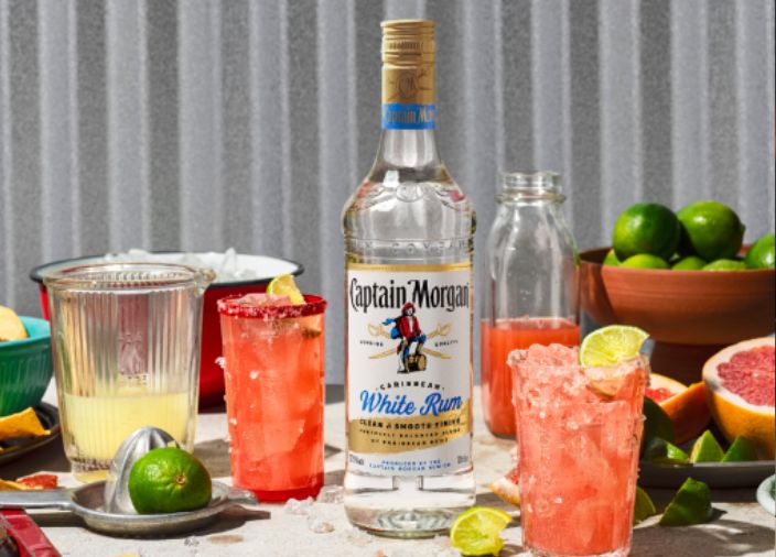 Botella de ron blanco Captain Morgan sobre la mesa rodeada de una variedad de alimentos y bebidas sobre fondo blanco.