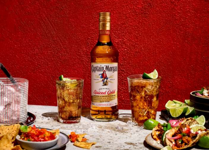 Flasche Captain Morgan Original Spiced Gold auf einem Tisch umgeben von verschiedenen Speisen und Getränken vor rotem Hintergrund