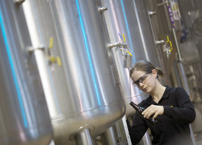 Female Diageo staff member wearing safety goggles, holding a brown beer bottle sampling from brewery fermentation tanks.