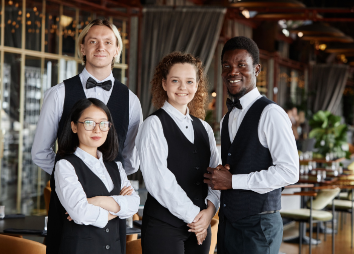 Equipe diversificada de quatro garçons sorrindo para a foto em um restaurante. Eles vestem uniformes clássicos com camisa branca e colete preto, dois com gravata borboleta. O elegante salão com mesas aparece desfocado ao fundo.