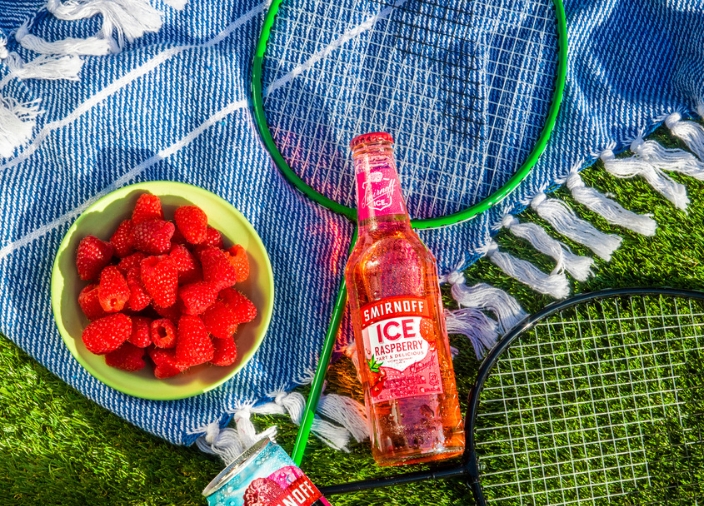 Botella de Smirnoff Ice Raspberry recostada sobre una manta sobre el césped con un plato de frambuesas al lado