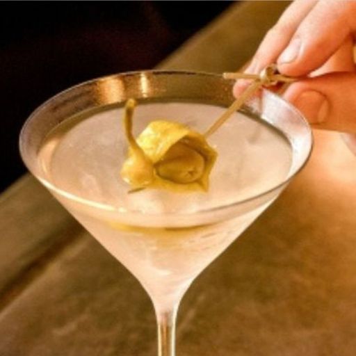 A martini drink with a bartender’s hand adding a green pickle garnish.