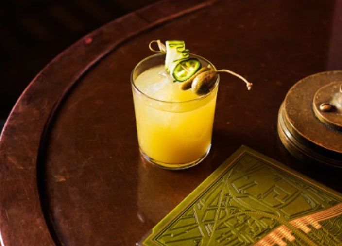 A yellow cocktail with a caperberry and cucumber garnish.