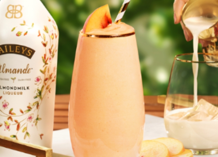 Una botella de Baileys Almande, con un Baileys Almande Peach Smoothie sin lactosa y un vaso con hielo en el que están sirviendo Baileys Almande.