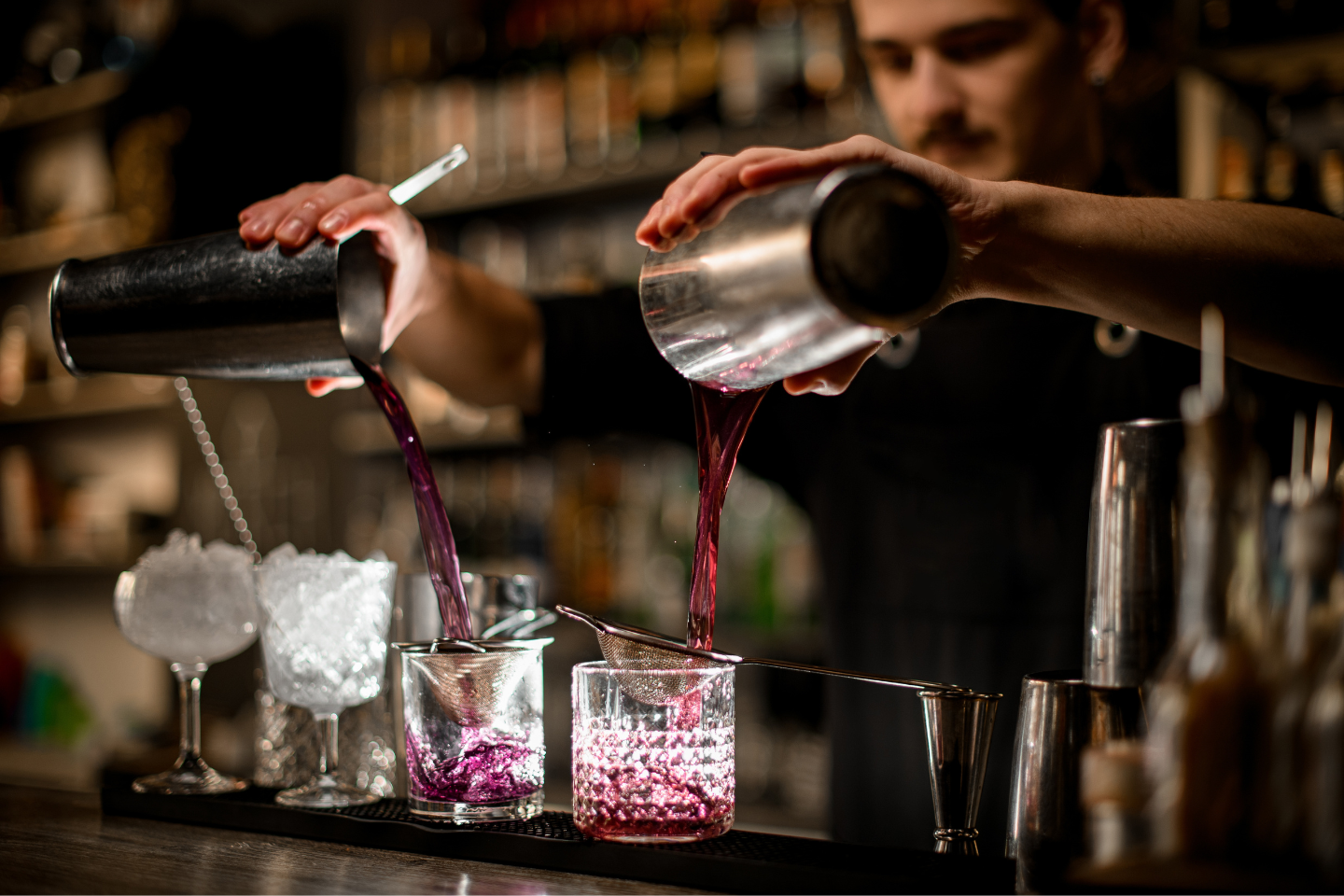 Mãos de bartender despejam simultaneamente um líquido roxo vibrante de duas coqueteleiras de prata em dois copos com gelo e um coador, em um ambiente de bar escuro.