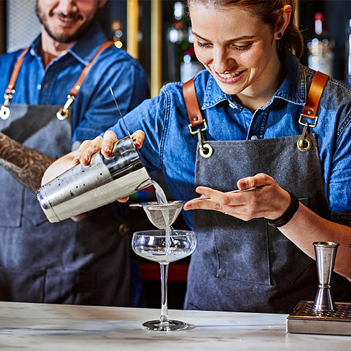 Ein weiblicher DBA-Barkeeper gießt einen Cocktail durch ein Sieb in ein klassisches Coupé-Cocktailglas.