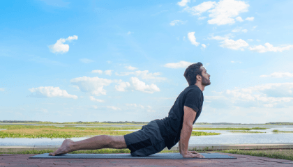 Man performing yoga pose outdoors on a mat with a scenic background and a blue sky with scattered clouds.