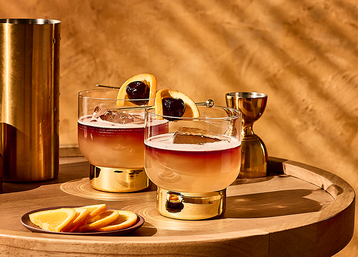 Dos copas de Whiskey Sours a base de Harper Cabernet junto a un platillo con rodajas de naranja, un jigger y un enfriador de vino sobre una mesa redonda de madera.