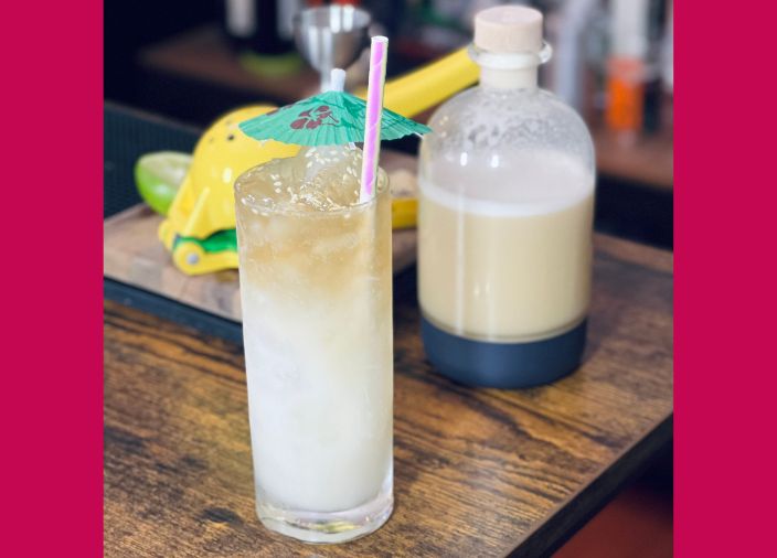 Ein weißer und hellbrauner Miso-Cocktail mit Eis, Sesamsamen und einem grünen Papierschirmchen neben einer Flasche Miso-Orangeat-Paste.