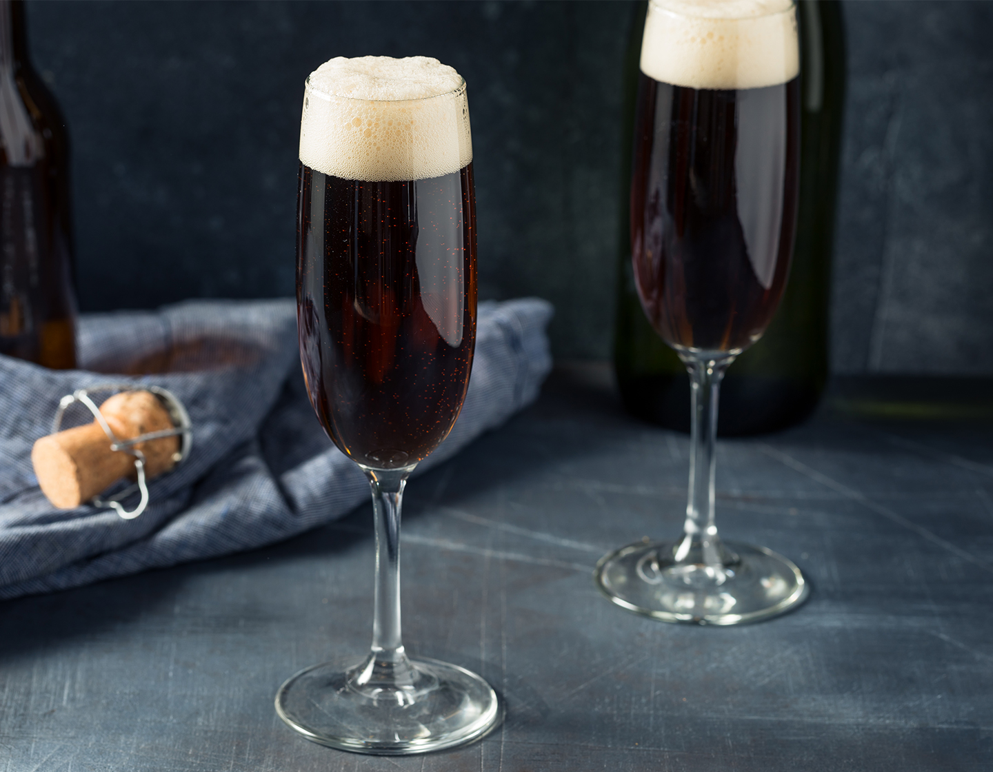 Dos cócteles Black Velvet en copas de vino espumoso escenificados con tela azul y el corcho de una botella de vino espumoso de fondo. 