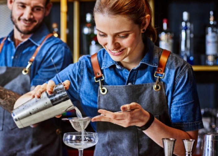 Female bartender strains cocktail into coupe glass with colleague.