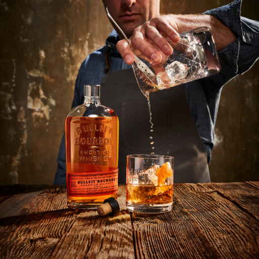 Un hombre cuela un cóctel en un vaso sobre una mesa junto a una botella abierta de whisky Bulleit.