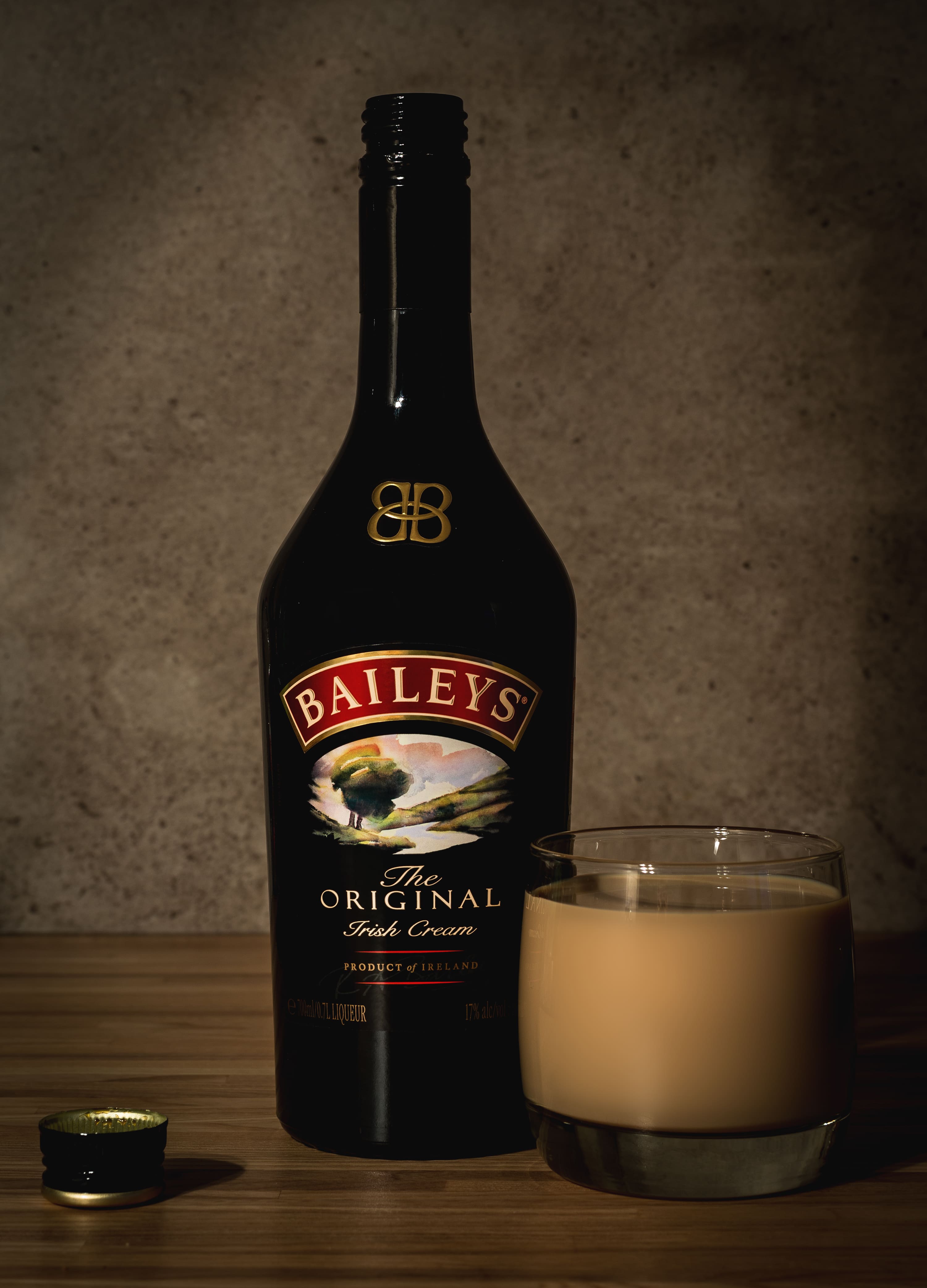 Garrafa de licor Baileys Original Irish Cream preta sobre uma mesa de madeira, ao lado de um copo baixo cheio da bebida cremosa de cor bege. A tampa da garrafa repousa sobre a mesa. Fundo de parede cinza texturizada com iluminação suave.
