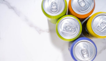 Vista superior de cinco latas de bebida coloridas em tons de verde, laranja, amarelo e roxo. As latas de alumínio estão fechadas, cobertas por gotas de condensação e agrupadas à direita sobre fundo de mármore branco.