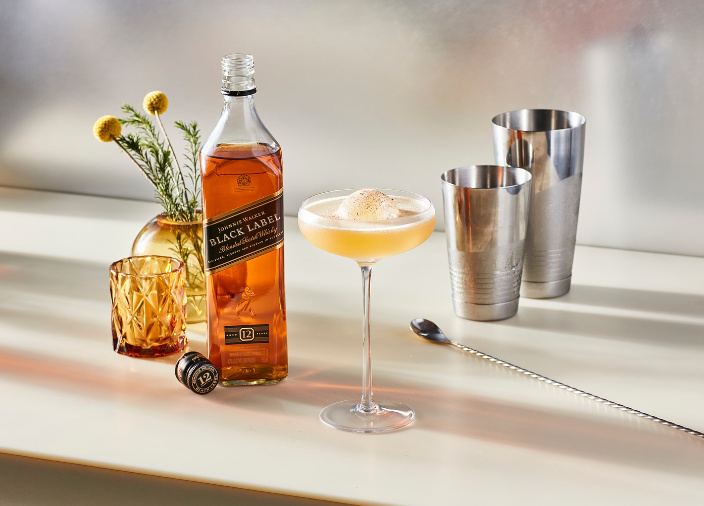 Cóctel en una copa tipo coupé con hielo, rodeado de una botella de Johnnie Walker Black Label Whisky, una coctelera y una cucharilla de bar.