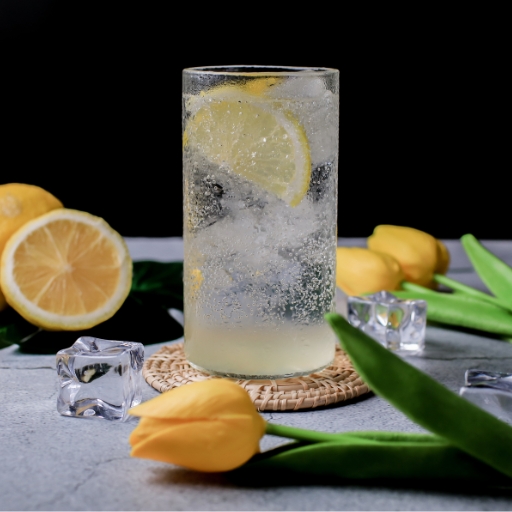 Bebida fría en un vaso transparente con rodajas de limón y hielo, colocada sobre una mesa con limones, hielo y flores amarillas.