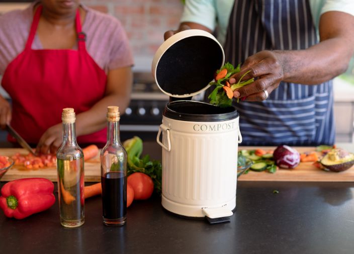 Putting fresh waste in a compost bin in front of people cutting veg on a chopping board.