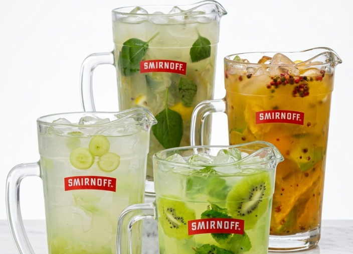 Quatro jarras de vidro com logo vermelho da Smirnoff sobre bancada clara. Cada uma contém um coquetel refrescante com gelo e ingredientes diferentes: uvas verdes fatiadas, kiwi com hortelã, maracujá e limão com folhas verdes. Fundo branco.