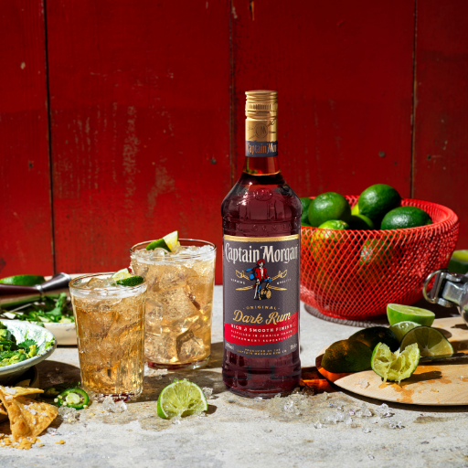 Bottle of Captain Morgan Dark Rum on table surrounded by assorted food and drink against red background