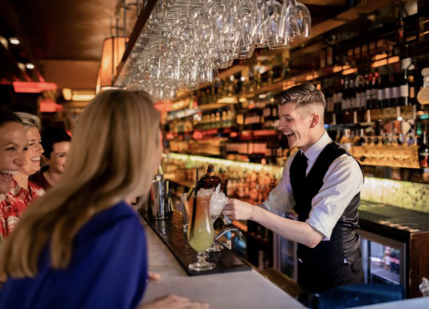 Ein Barkeeper lacht und unterhält sich mit Gästen an der Bar, während er ein Getränk einschenkt.