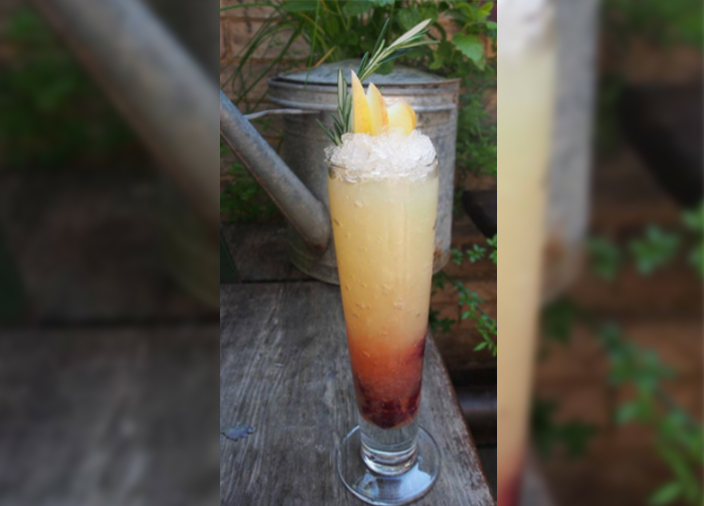 A homemade cocktail with red liquid at the bottom and yellow and white at the top, garnished with sliced apple, in a sling style cocktail glass.