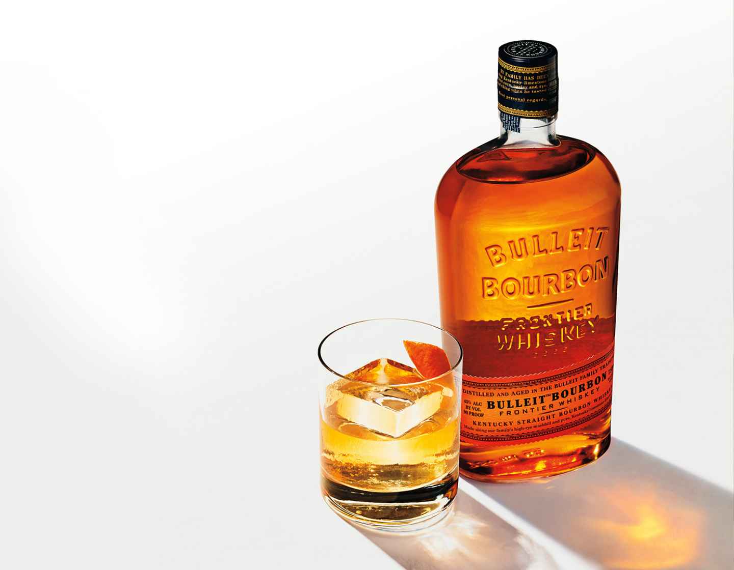 Uma garrafa de Bulleit Bourbon em uma mesa branca ao lado de um copo cheio de Bulleit e um grande cubo de gelo. Sombras brilhantes são projetadas no canto inferior direito.