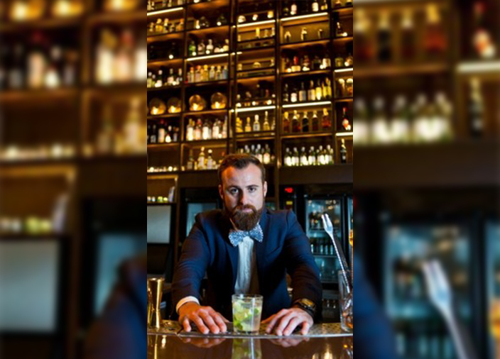 Tai Barbin, elegantemente vestido con chaqueta de traje y corbata de moño, presentando un cóctel Caipirinha.