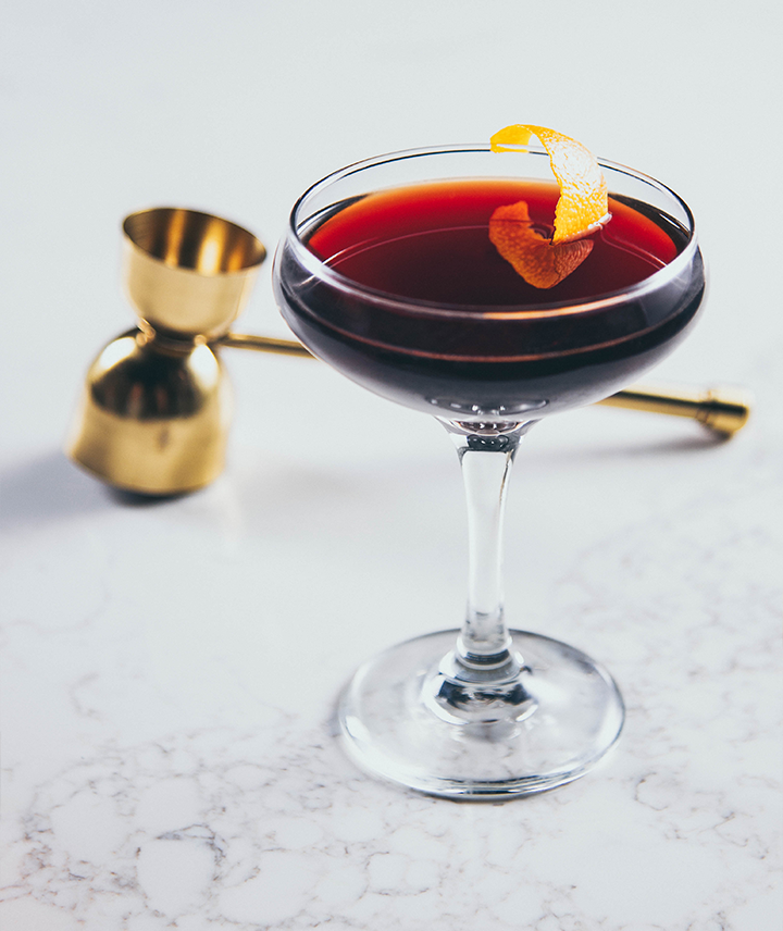Cocktail in a coupe glass with orange garnish and gold jigger