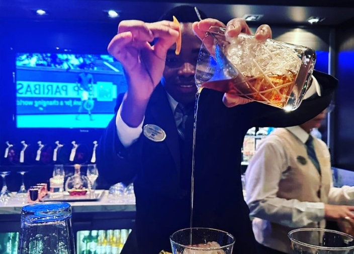 A man behind a bar pouring liquid from a jug with ice and a strainer into a glass at height, holding an orange garnish in the other hand.