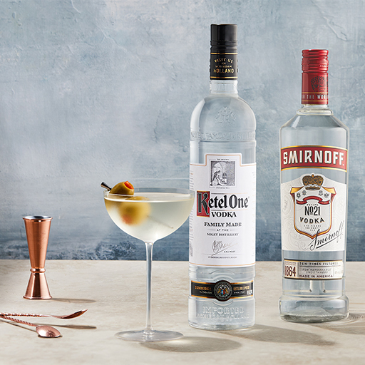 Una botella de vodka Ketel One y vodka Smirnoff sobre una mesa, junto a un vaso de cóctel clásico que contiene una bebida fría y una guarnición de dos aceitunas. Un medidor y una cucharilla mezcladora de color oro rosa están colocados sobre la mesa.