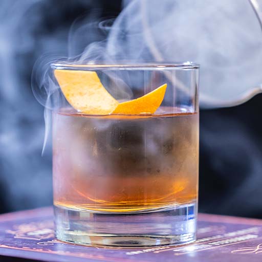Un vaso transparente lleno de líquido y una cáscara de naranja rodeada de humo.