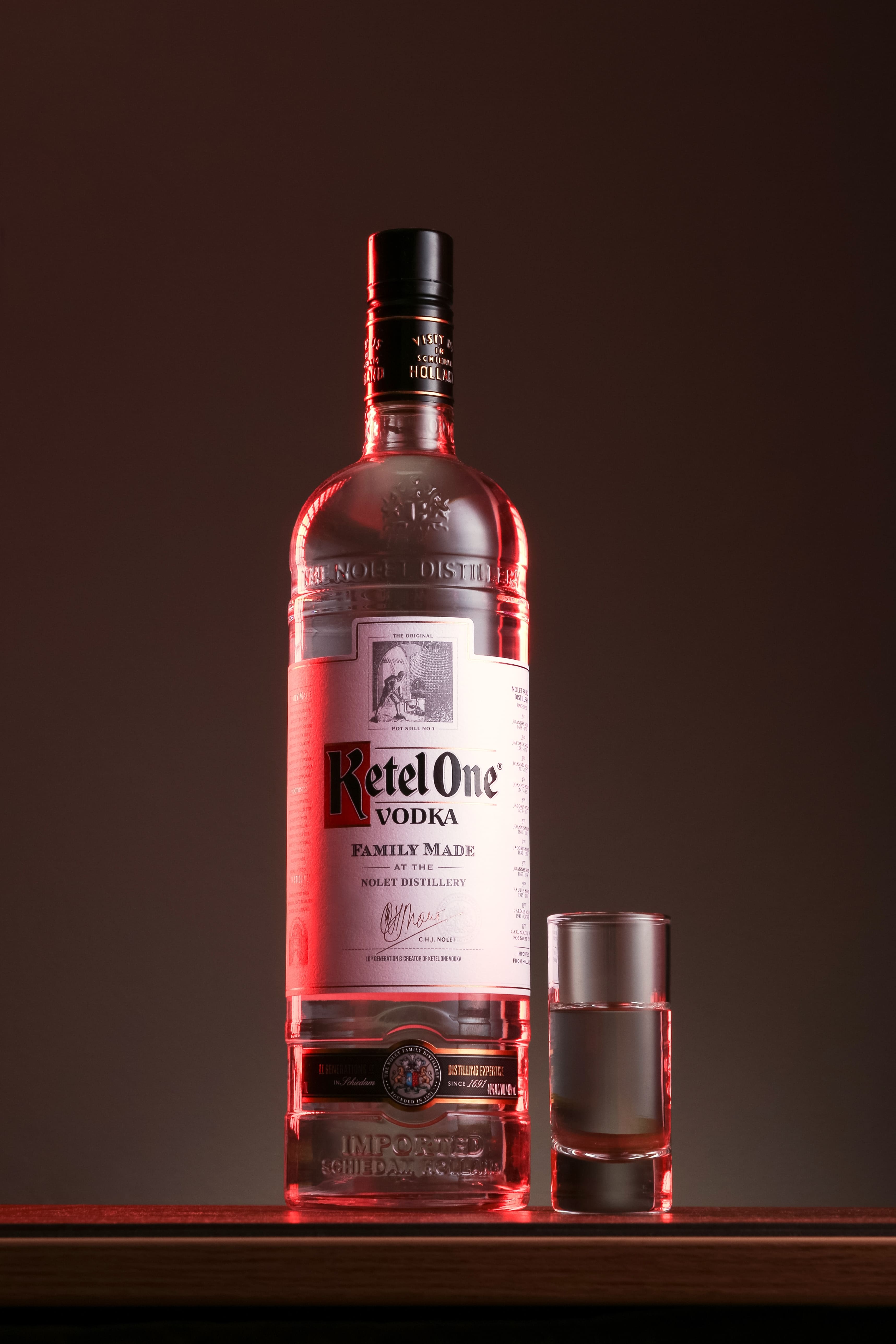 Garrafa de vodka Ketel One com rótulo branco e letras vermelhas, ao lado de um copo de shot cheio de líquido transparente. Iluminação traseira vermelha cria um brilho rosado nos contornos da garrafa e do copo. Fundo escuro.