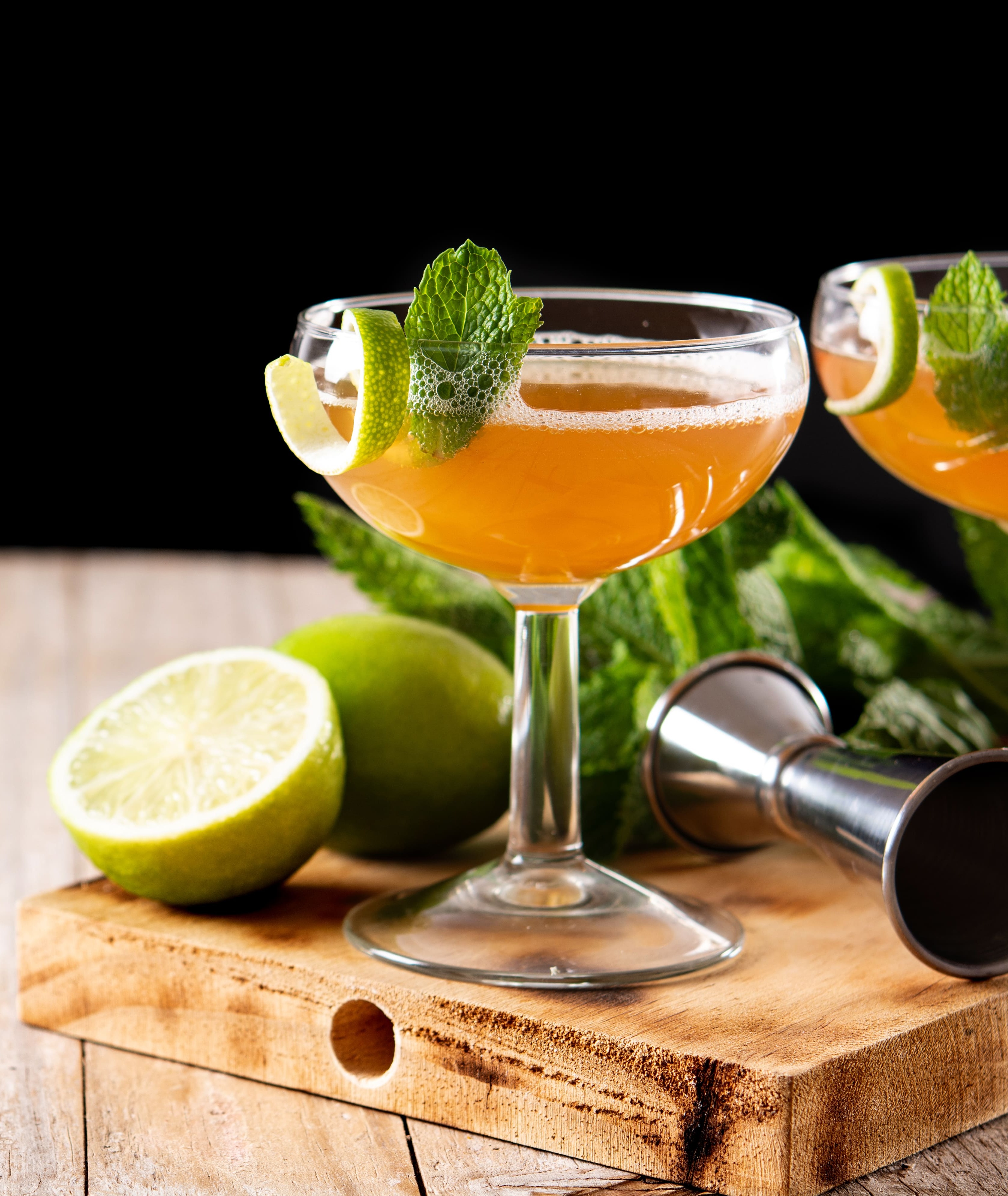 Dois coquetéis de cor laranja em taças coupe, decorados com hortelã fresca e casca de limão, sobre uma tábua de madeira rústica. Ao lado, limões cortados e um dosador de metal prateado. O fundo é preto, contrastando com a madeira clara da mesa.
