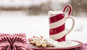 Uma caneca temática natalina em cima de um pires recheado de guloseimas, com neve ao fundo