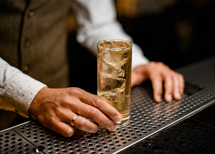 Dos manos sosteniendo un highball con cubitos de hielo y líquido de color ámbar