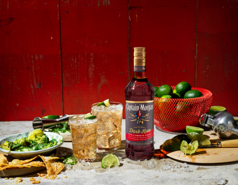Botella de Captain Morgan Dark Rum sobre una mesa rodeada de comida y bebida variada frente a un fondo rojo.