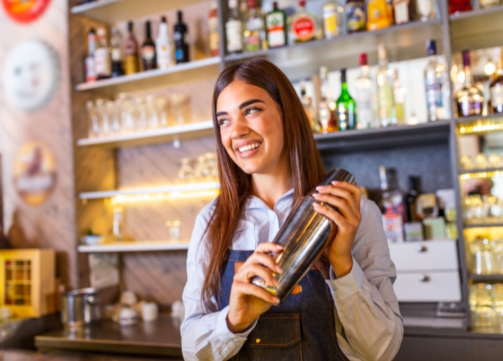 Uma mulher com longos cabelos escuros sacudindo uma coqueteleira atrás de um bar, sorrindo.